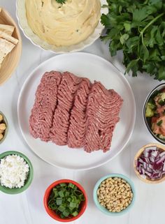 meat and vegetables are arranged on a white table with bowls of salad, pita bread, hummuss, olives