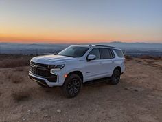 a white suv parked on top of a dirt hill next to the ocean at sunset