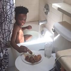 a young boy sitting in a bathtub with food on a plate next to him