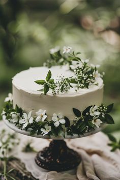 a white cake with flowers and greenery on top sitting on a table in the woods