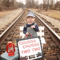 a baby holding a sign that says chugga chuga two two on the train tracks