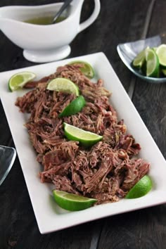 a white plate topped with shredded beef and limes next to two cups of tea