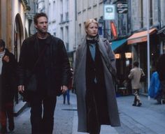 the man and woman are walking down the street in front of some buildings, one is wearing a long coat