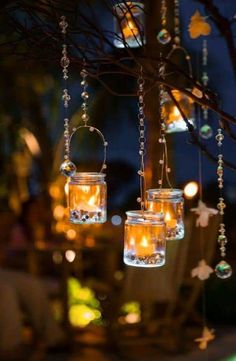 three glass jars filled with candles hanging from a tree branch in front of some lights