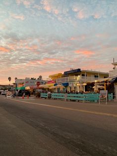 the sun is setting on an empty street in front of some buildings and shops with people walking by