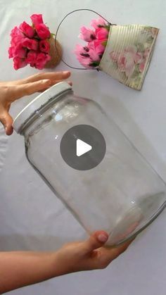 a person holding a glass jar with pink flowers in it on top of a table