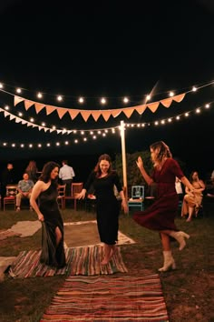 two women in dresses are dancing on a rug with lights strung over the ground and people sitting around