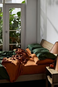 an unmade bed with green and orange sheets in front of a window next to a chair