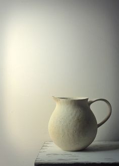 a white vase sitting on top of a wooden table next to a wall with a light behind it