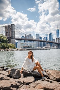 a woman sitting on rocks near the water in front of a bridge and cityscape