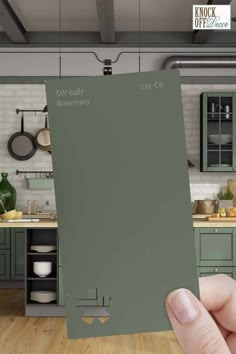 a person holding up a green paint swatch in a kitchen with grey cabinets and counters