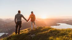 two people holding hands while standing on top of a hill with the sun setting in the background