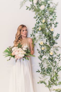 a woman in a white dress holding a bouquet and standing next to a wall with flowers