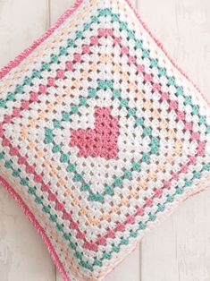 a crocheted square pillow on a white wooden surface with pink and green accents