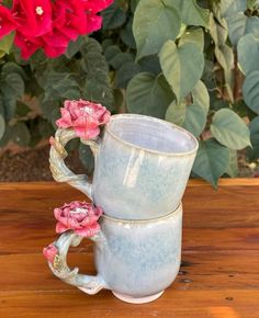 two coffee mugs sitting on top of a wooden table next to red and green flowers