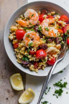 a white bowl filled with shrimp, rice and tomatoes on top of a table next to lemon wedges