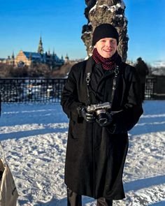 a man standing in the snow with a camera on his shoulder and wearing a black coat