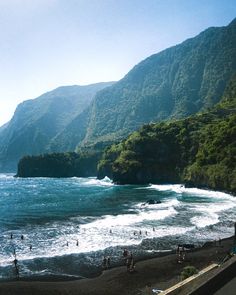 many people are on the beach near some mountains and water, while others swim in the ocean