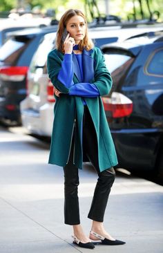 a woman is standing on the sidewalk talking on her cell phone while wearing black pants and a green coat