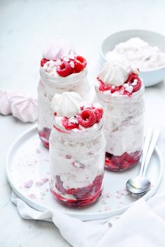 three jars filled with whipped cream and raspberries on a white plate next to two spoons