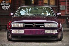 the front end of a purple car parked in front of a brick building with an emblem on it