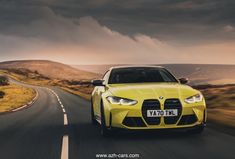 a yellow car driving down the road with dark clouds in the sky behind it and grass on both sides