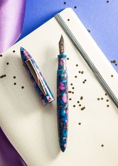 two pens sitting on top of a notepad next to a purple and blue cloth