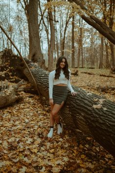 a woman leaning against a fallen tree in the woods with leaves on the ground and trees behind her