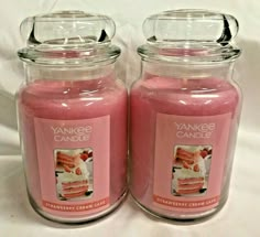 two yankee candles sitting next to each other on a white tablecloth covered surface, one is pink and the other has an image of a piece of cake