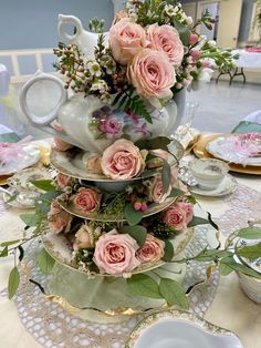 three tiered cake stand with pink roses and greenery on top, surrounded by tea cups and saucers