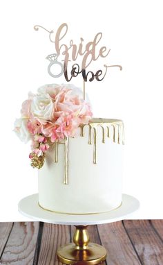 a white and gold wedding cake with pink flowers on the top is sitting on a wooden table