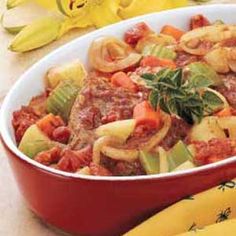 a red dish filled with pasta and meat next to yellow flowers on a tablecloth