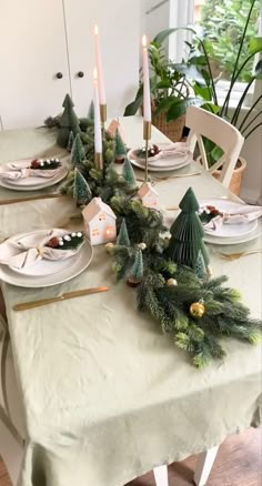 the table is set for christmas dinner with pine trees and candles on it, along with other holiday decorations