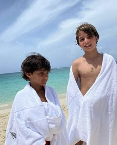 two young men standing on top of a sandy beach next to the ocean with towels draped around them