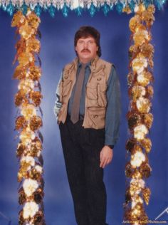 a man with a moustache standing in front of a blue background wearing a vest and tie