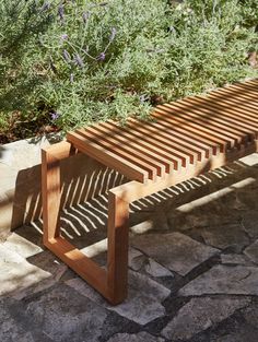 a wooden bench sitting on top of a stone floor next to bushes and flowers in the sun