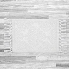 a white placemat with tassels on top of a wooden floor in front of a gray wall