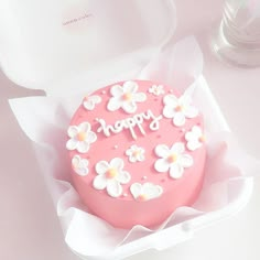 a pink birthday cake with white flowers on top in a paper box next to a glass container