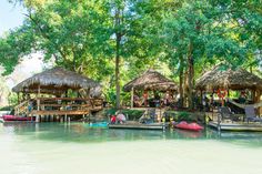 some people are sitting on the dock in front of thatched huts and canoes