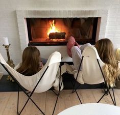 two women sitting in chairs facing an open fire with their feet up on the table