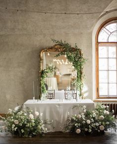 a table with white flowers and greenery on it in front of a large mirror