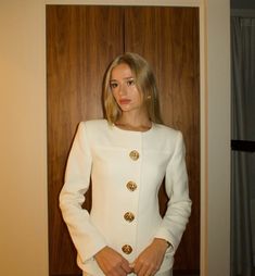 a woman standing in front of a wooden door wearing a white suit and gold earrings