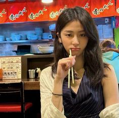 a woman holding chopsticks in her mouth while sitting at a table with food on it