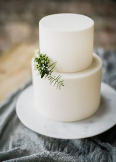 two tiered white cake with greenery on top sitting on a table next to a gray blanket