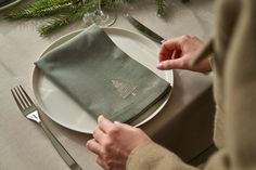 a person setting a place setting on a table with silverware, napkins and evergreen branches