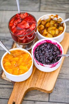 four bowls of fruit and berries on a cutting board with spoons in each bowl