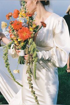 a woman in a white dress holding a bouquet of orange and yellow flowers with other people behind her