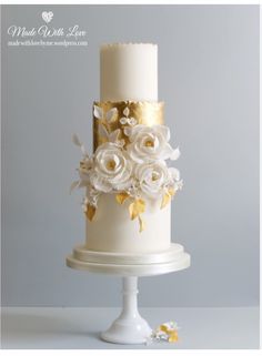 a three tiered white and gold wedding cake with flowers on the top, sitting on a pedestal