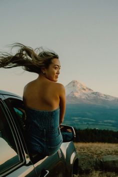 a woman standing on the hood of a car with her hair in the wind and mountains in the background