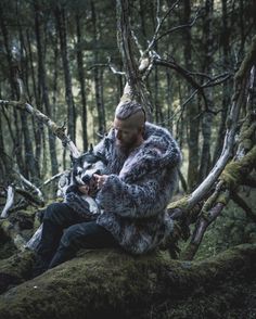 a man in a fur coat sitting on a mossy rock with his dog and looking at the camera